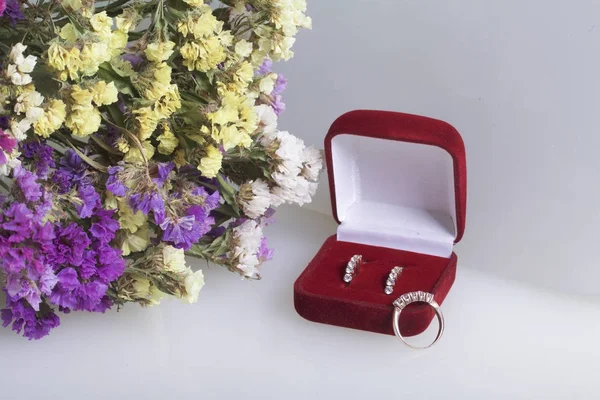 A bouquet of dried flowers lies on a white surface. Nearby is an open velvet box with a gift. Earrings and a ring for your beloved.