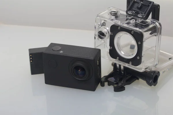 The action camera lies on a white surface. Near the box for underwater shooting and a spare battery.