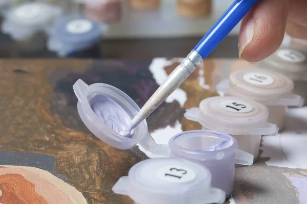 Painting on canvas by numbers. A woman is holding a brush and drawing it. Numbered containers with paints and brushes on the canvas background.