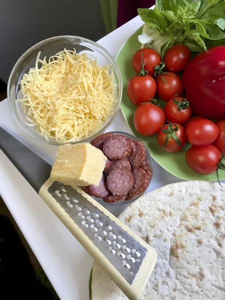 Preparación de pizza. Los ingredientes necesarios están sobre la mesa: queso, salchichas, pizza base y verduras . —  Fotos de Stock
