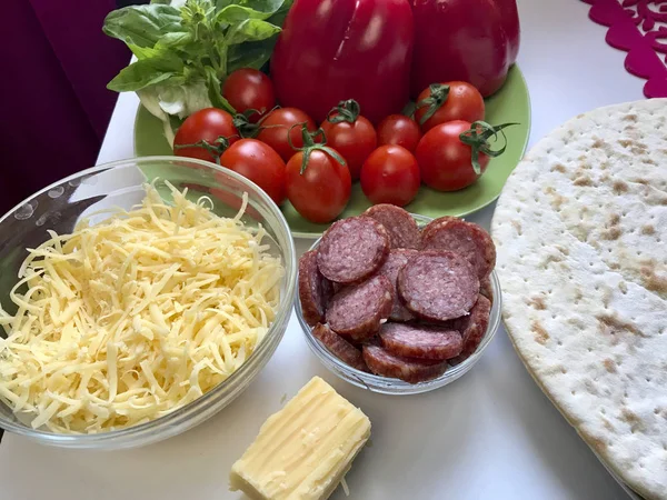 Příprava pizzy. Na stole jsou potřebné ingredience: sýr, klobása, pizza základny a zeleniny. — Stock fotografie