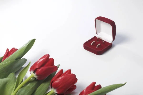 Dárky pro blízké. Kytice červených tulipánů je rozptýlena na bílém povrchu. Nedaleko je krabice červeného sametu s prsten a náušnice. — Stock fotografie