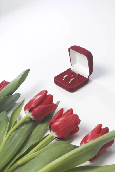 Dárky pro blízké. Kytice červených tulipánů je rozptýlena na bílém povrchu. Nedaleko je krabice červeného sametu s prsten a náušnice. — Stock fotografie