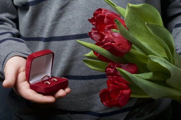 Presentes para entes queridos. Os homens estão segurando um buquê de tulipas vermelhas na mão. Por outro lado, uma caixa de veludo aberto de cor vermelha, em que um anel e brincos . — Fotografia de Stock