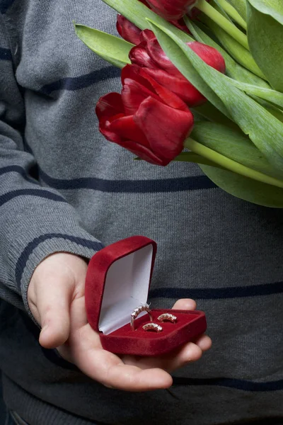Dárky pro blízké. Muži se v ruce drží kytici červené tulipány. Na druhou stranu, otevřené velvet box červené barvy, v němž prsten a náušnice. — Stock fotografie