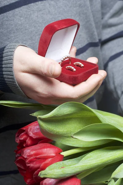 Presentes para entes queridos. Os homens estão segurando um buquê de tulipas vermelhas na mão. Por outro lado, uma caixa de veludo aberto de cor vermelha, em que um anel e brincos . — Fotografia de Stock