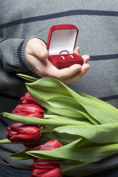 Dárky pro blízké. Muži se v ruce drží kytici červené tulipány. Na druhou stranu, otevřené velvet box červené barvy, v němž prsten a náušnice. — Stock fotografie