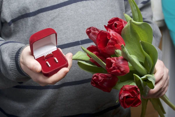 Presentes para entes queridos. Os homens estão segurando um buquê de tulipas vermelhas na mão. Por outro lado, uma caixa de veludo aberto de cor vermelha, em que um anel e brincos . — Fotografia de Stock