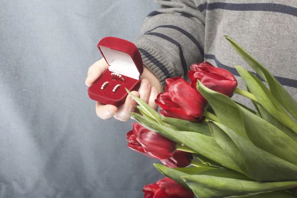 Presentes para entes queridos. Os homens estão segurando um buquê de tulipas vermelhas na mão. Por outro lado, uma caixa de veludo aberto de cor vermelha, em que um anel e brincos . — Fotografia de Stock