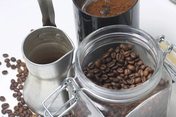 Cezve se tient sur une surface blanche. À côté d'un pot en verre avec des grains de café et un moulin à café avec du café moulu . — Photo