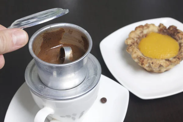 Vietnamese coffee maker is equipped on a cup. It is filled with ground coffee and pour boiling water. There is a cake on the saucer. The man lifted the lid of the brewer.