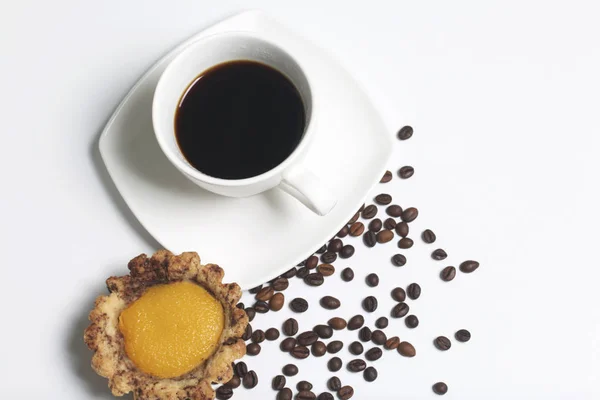 En la superficie blanca de la mesa hay una taza de café fragante en un platillo. Hay un pastel cerca. Siguiente granos de café dispersos . — Foto de Stock