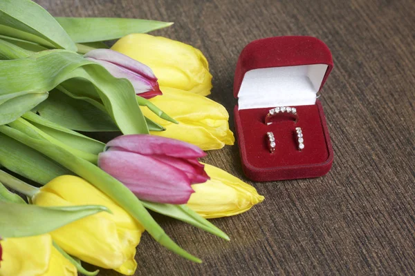 Presentes para entes queridos. Um buquê de tulipas amarelas e rosa é espalhado em uma superfície escura. Nas proximidades é uma caixa de veludo aberta de cor vermelha com jóias de ouro . — Fotografia de Stock