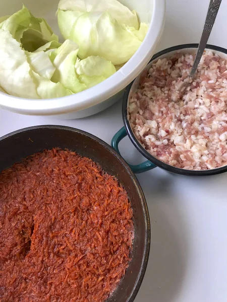 En la mesa están los ingredientes para cocinar rollos de col. Zanahorias infladas en una sartén, carne picada en un recipiente y hojas de col en un recipiente . — Foto de Stock