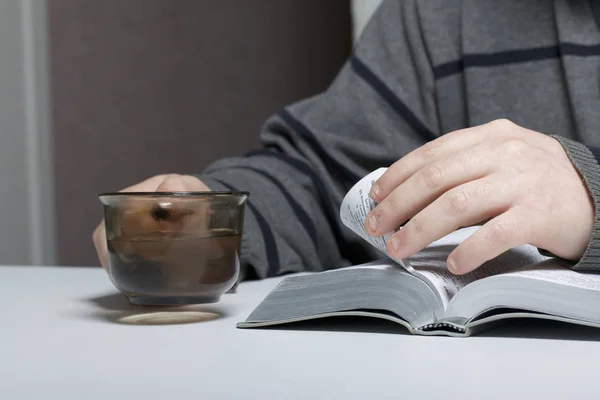 Un joven lee la Biblia con una rebanada de plata sentada a la mesa. Una taza con agua está al lado. . — Foto de Stock