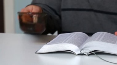 Bir adam İncil okur, sayfalar arasında çevirir ve kupa suyu içer.