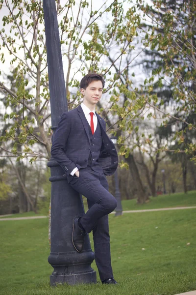 Um jovem, um adolescente, num fato clássico. Contando com uma lanterna vintage no parque. Põe as mãos nos bolsos. . — Fotografia de Stock
