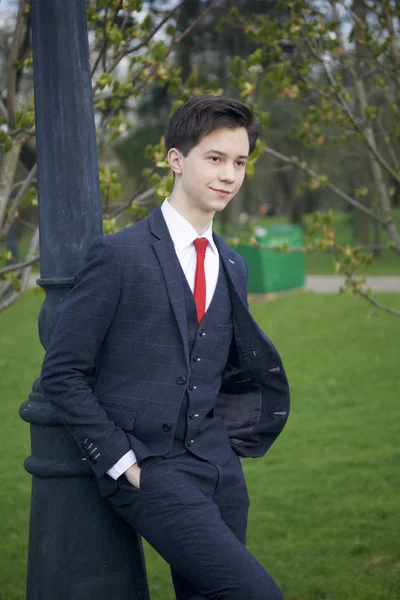 Un joven, un adolescente, con un traje clásico. Confiando en una linterna vintage en el parque. Pon las manos en los bolsillos. . —  Fotos de Stock