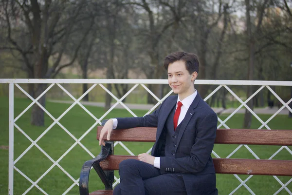 Een jonge man, tiener, in een klassieke pak. Zit op een vintage Bank in een park voorjaarsbijeenkomst. — Stockfoto