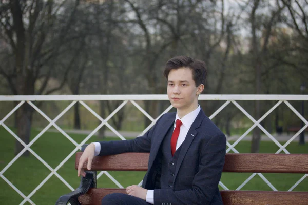 Молодой человек, подросток, в классическом костюме. Сидит на винтажной скамейке в весеннем парке . — стоковое фото