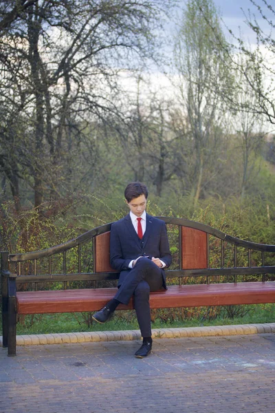 Um jovem, um adolescente, num fato clássico. Senta-se em um banco vintage em um parque de primavera. Ele segura um smartphone em suas mãos e olha para sua tela . — Fotografia de Stock