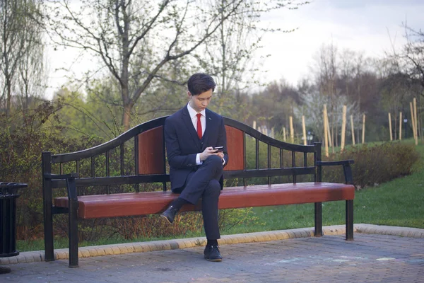 Un jeune homme, un adolescent, en costume classique. Assis sur un banc vintage dans un parc de printemps. Il tient un smartphone dans ses mains et regarde son écran . — Photo