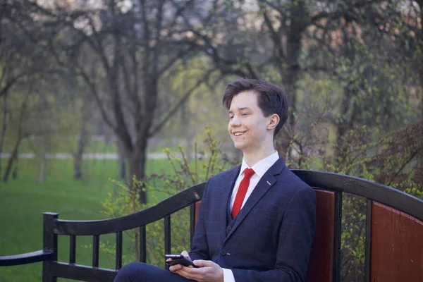 Een jonge man, tiener, in een klassieke pak. Zit op een vintage Bank in een park voorjaarsbijeenkomst. Hij houdt van een smartphone in zijn handen en gelukkig kijkt. — Stockfoto