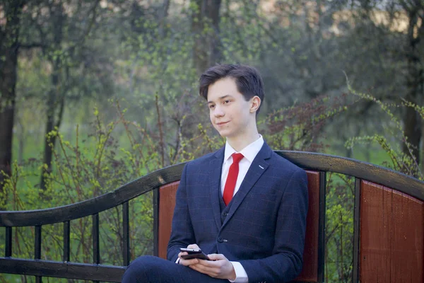 Młody człowiek, nastolatek, w klasycznym garniturze. Siedzi na ławce vintage w parku wiosny. Posiada smartfona w jego ręce i zamyśleniu patrzy w dal. — Zdjęcie stockowe
