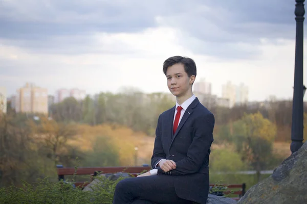 Młody człowiek, nastolatek, w klasycznym garniturze. On siedzi na duży głaz w parku wiosny, ręce splecione na kolanach. — Zdjęcie stockowe