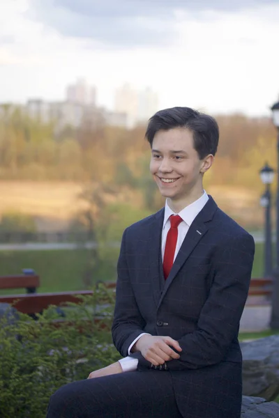 Un jeune homme, un adolescent, en costume classique. Il est assis sur un grand rocher dans un parc de printemps, les mains jointes sur ses genoux . — Photo