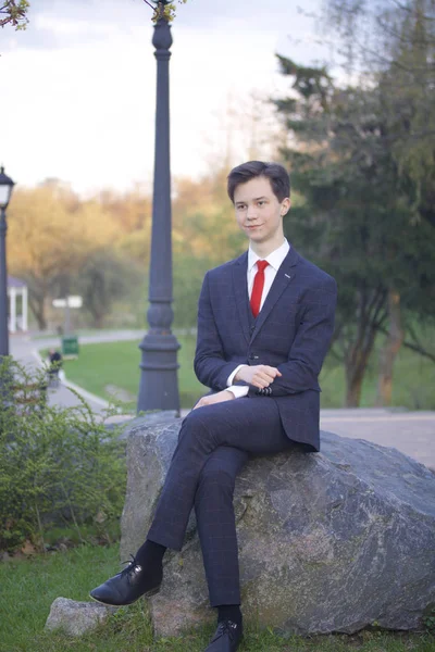Un jeune homme, un adolescent, en costume classique. Il est assis sur un grand rocher dans un parc de printemps, les mains jointes sur ses genoux . — Photo