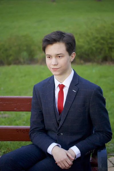 Ein junger Mann, ein Teenager, in einem klassischen Anzug. sitzt auf einer Vintage-Bank im Frühlingspark. — Stockfoto