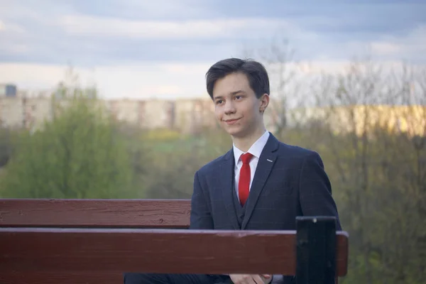 Un joven, un adolescente, con un traje clásico. Se sienta en un banco vintage en un parque de primavera . —  Fotos de Stock