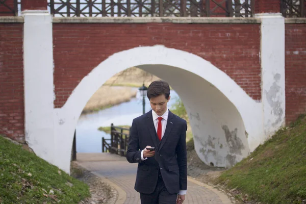 Un jeune homme, un adolescent, en costume classique. Il tient un smartphone dans ses mains, et le regarde en déplacement . — Photo