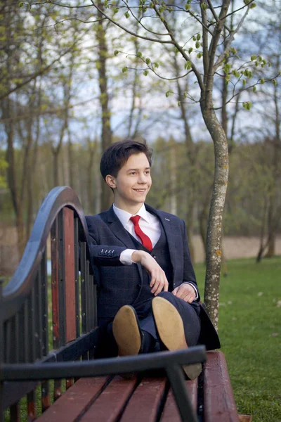 Um jovem, um adolescente, num fato clássico. Senta-se em um banco vintage em um parque de primavera . — Fotografia de Stock