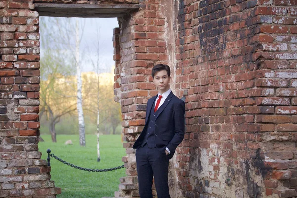 Молодой человек, подросток, в классическом костюме. Глупо стоит напротив старой стены из красного кирпича, засовывая руки в карманы . — стоковое фото