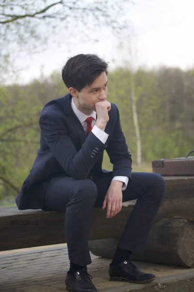 Un jeune homme, un adolescent, en costume classique. Assis sur un banc en bois dans un parc de printemps. Il y réfléchit, étendant sa tête avec sa main . — Photo