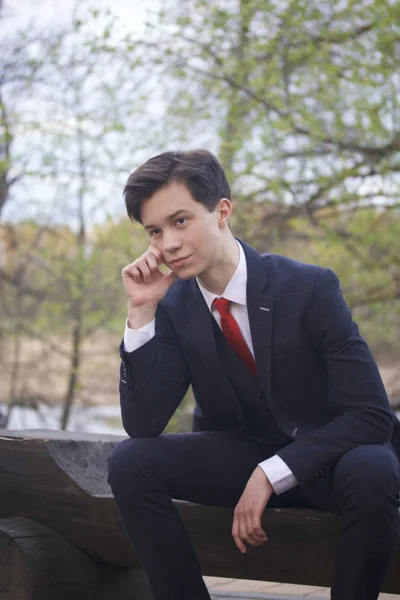 Un jeune homme, un adolescent, en costume classique. Assis sur un banc en bois dans un parc de printemps. Il y réfléchit, étendant sa tête avec sa main . — Photo