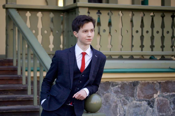 Молодой человек, подросток, в классическом костюме. Он стоит, опираясь на перила деревянной лестницы . — стоковое фото