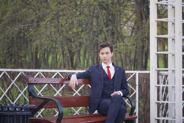 Un jeune homme, un adolescent, en costume classique. Assis sur un banc vintage dans un parc de printemps . — Photo