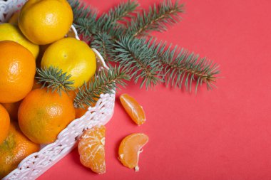 Hasır sepette mandalina. Sepetin yanında soyulmuş mandalina var. Görünür mandalina dilimleri ve derisi. Yeşil ladin dalları. Mercan renginin arka planına karşı.