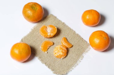 Mandarinler beyaz bir yüzeye dizilmiştir. Yanlarındaki çarşaf parçasının üzerinde mandalina kabuğu var. Görünür mandalina dilimleri. Beyaz arkaplanda.