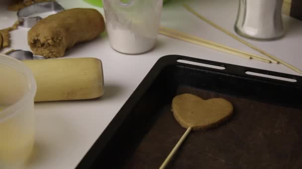 La donna sta preparando dei biscotti di pan di zenzero. Mantiene un cookie vuoto e lo corregge. Un pezzo di pasta a forma di cuore infilato su un bastoncino giace su una teglia da forno . — Video Stock