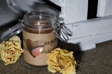 Kağıt parçalarıyla sarılmış cam kavanoz. Kâğıda kazınmış bir kalp. Çarşaf örtüsünün üzerinde. Bankada bir mum yanıyor. Yakınlarda kurumuş gül tomurcukları ve tahta bir kutu..