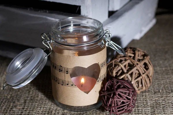 Kağıt parçalarıyla sarılmış cam kavanoz. Kâğıda kazınmış bir kalp. Çarşaf örtüsünün üzerinde. Bankada bir mum yanıyor. Yanında rattan topları ve tahta bir kutu..