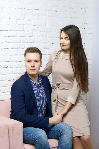 Ένας νεαρός κάθεται σε μια καρέκλα και κρατά τη γυναίκα του από το χέρι. Μια σύζυγος στέκεται κοντά και αγκαλιάζει το λαιμό του. — Φωτογραφία Αρχείου