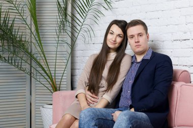 Genç bir aile kanepede oturuyor. Şenlikli giyinmiş. Neşeli bir ruh hali. Yakınlarda saksıda bir palmiye ağacı var..