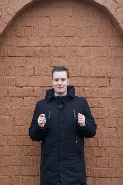 Siyah ceketli genç bir adam kahverengi tuğla bir duvarın arkasında poz veriyor. Önünde el ele tutuşuyor.. — Stok fotoğraf