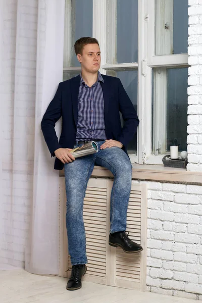 Jeune homme avec une expression arrogante. Assis près de la fenêtre sur le rebord de la fenêtre. Dans sa main tient un magazine tordu dans un tube . — Photo