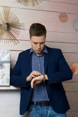 Düşünceli bakışlı genç bir adam kol saatinde saati izler. Dirsek bir rafta duruyor..